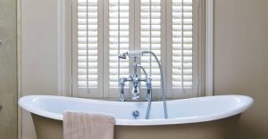 White wooden window plantation  shutters in traditional bathroom with free-standing bath in foreground.